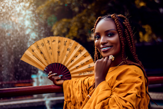 a woman with braids holding a fan in front of a fountain