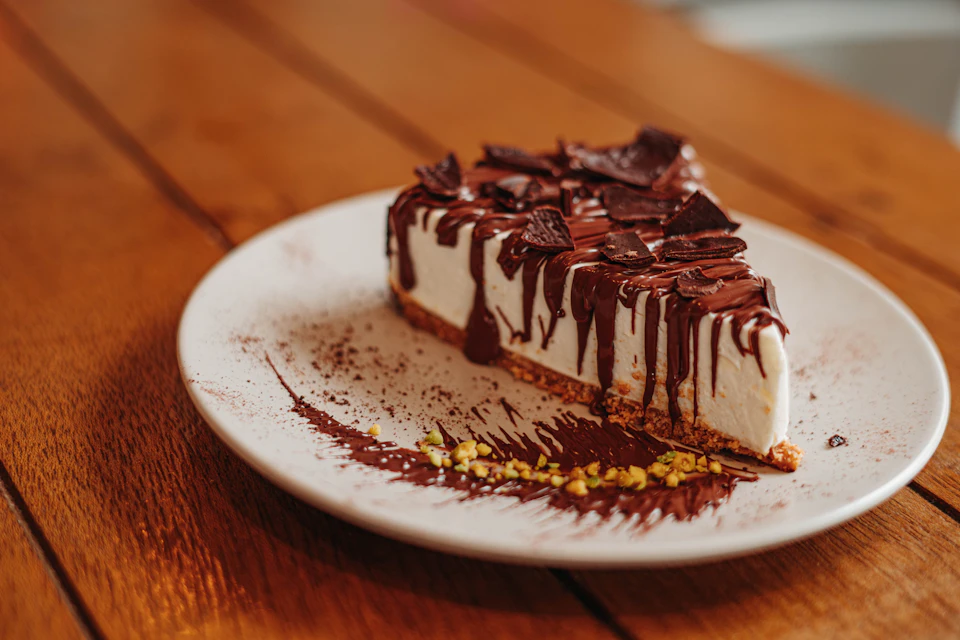 The image shows a slice of chocolate cake on a white plate, resting on a wooden table. The cake is covered in chocolate icing and topped with chocolate chips. The plate has a white rim around it, adding an elegant touch to the presentation.
