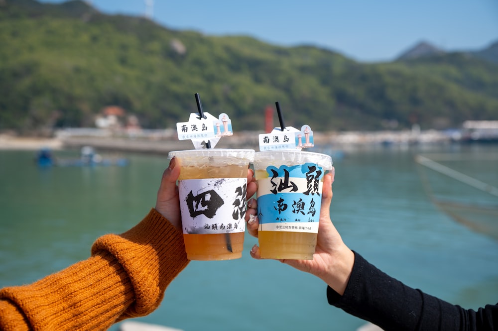 two people holding up cups with drinks in front of a body of water