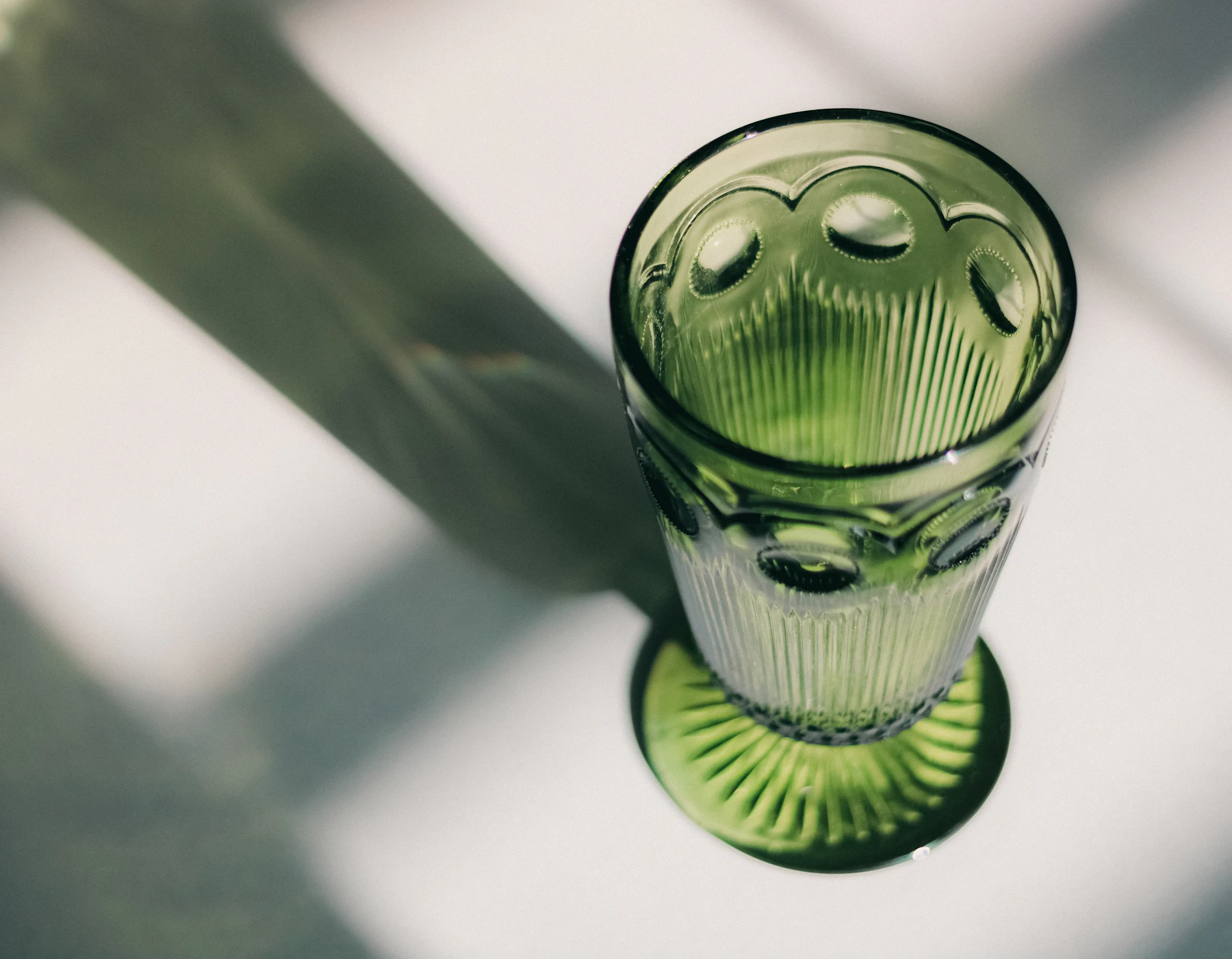 A green pressed-glass footed tumbler seen from above, casting a long shadow in window light