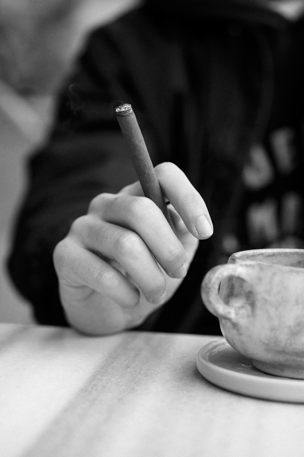a person sitting at a table with a cup and a cigarette