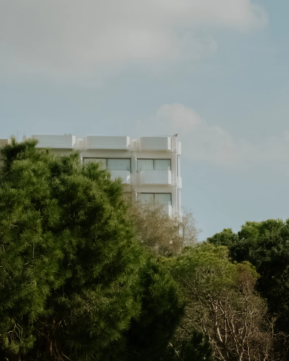 The image depicts a modern, white building with numerous windows and balconies situated amidst lush green trees. The building's design is sleek and contemporary, contrasting with the natural surroundings. The perspective of the photo is from below, looking up at the towering structure against a clear blue sky dotted with fluffy white clouds.