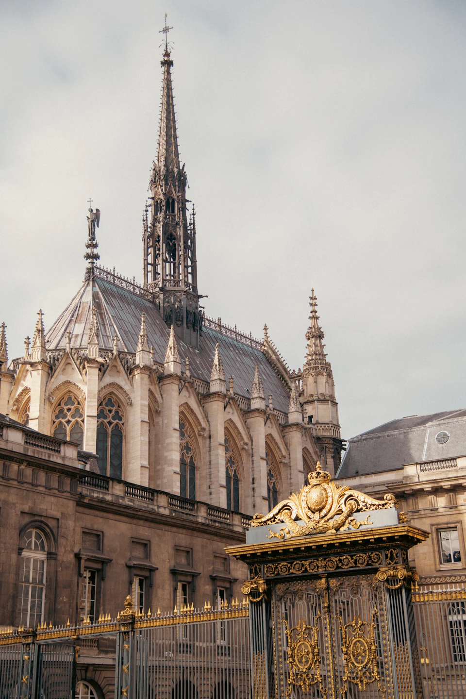 The image depicts a large, ornate building with a tall spire and a clock tower reaching towards the sky. The building is constructed from stone and features intricate carvings on its facade. A gate stands at the entrance to the building, which is adorned with gold accents that shimmer under the light.

In front of the building, there's a large, ornate gate with a gold-colored roof and a clock face on each side. The sky above is overcast, casting a soft light over the scene.