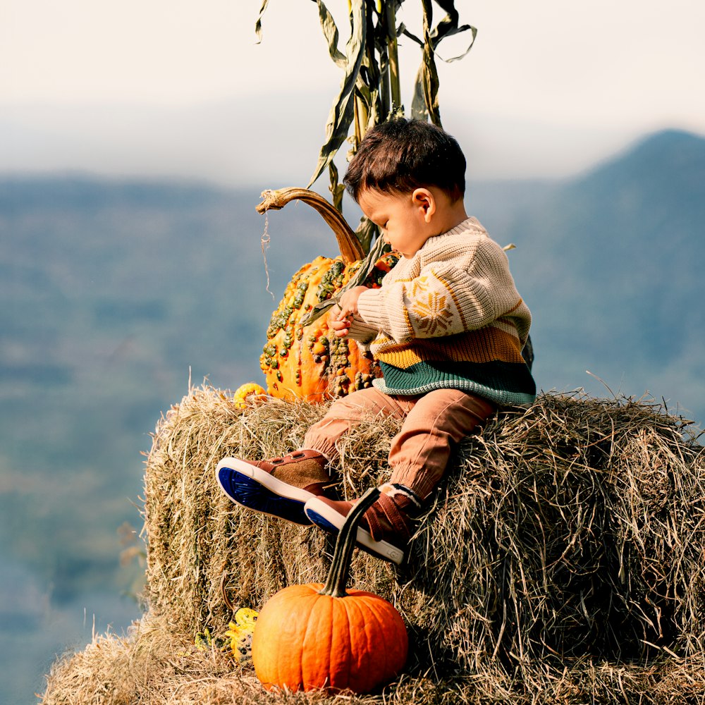 a young boy sitting on a bale of hay next to a pumpkin