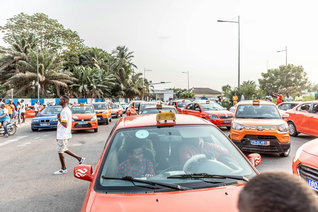 Des taxis orange à l’arrêt dans une rue bordée de palmiers à Abidjan, entourés de piétons