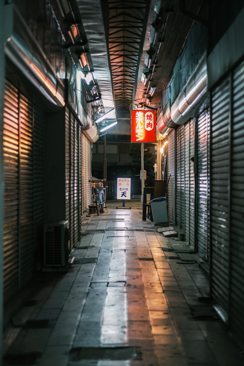 The image depicts a narrow alleyway with a row of shops on either side, their shutters closed and illuminated by warm yellow lights that cast long shadows across the cobblestone street. The buildings are made of stone or concrete, giving them an old-world charm despite being in a modern city setting. The perspective is from ground level, looking down the alleyway towards the end of it.