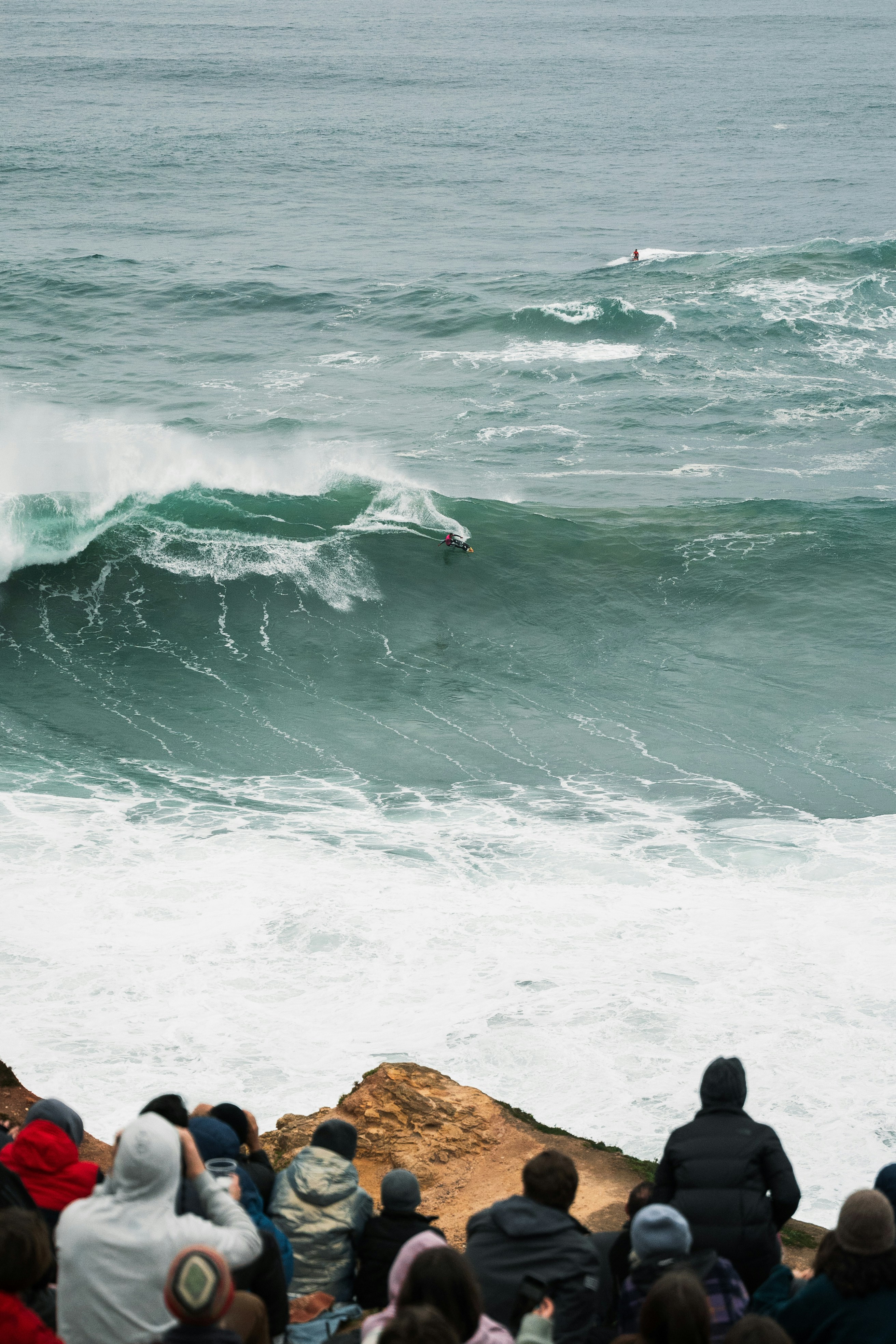 Spectators watching a surfer ride a wave from a clifftop viewpoint in Nazaré, Portugal
