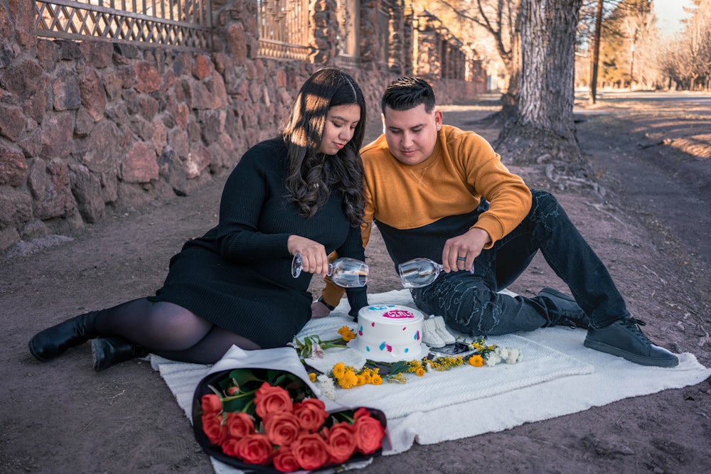 a man and woman sitting on a blanket cutting a cake