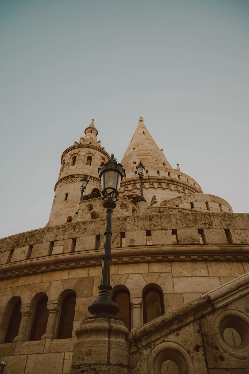 The image depicts a large stone building with multiple towers and spires, standing tall against a clear blue sky. The building is situated on a street corner, with a lamppost nearby. The perspective of the photo is from below, looking up at the imposing structure.