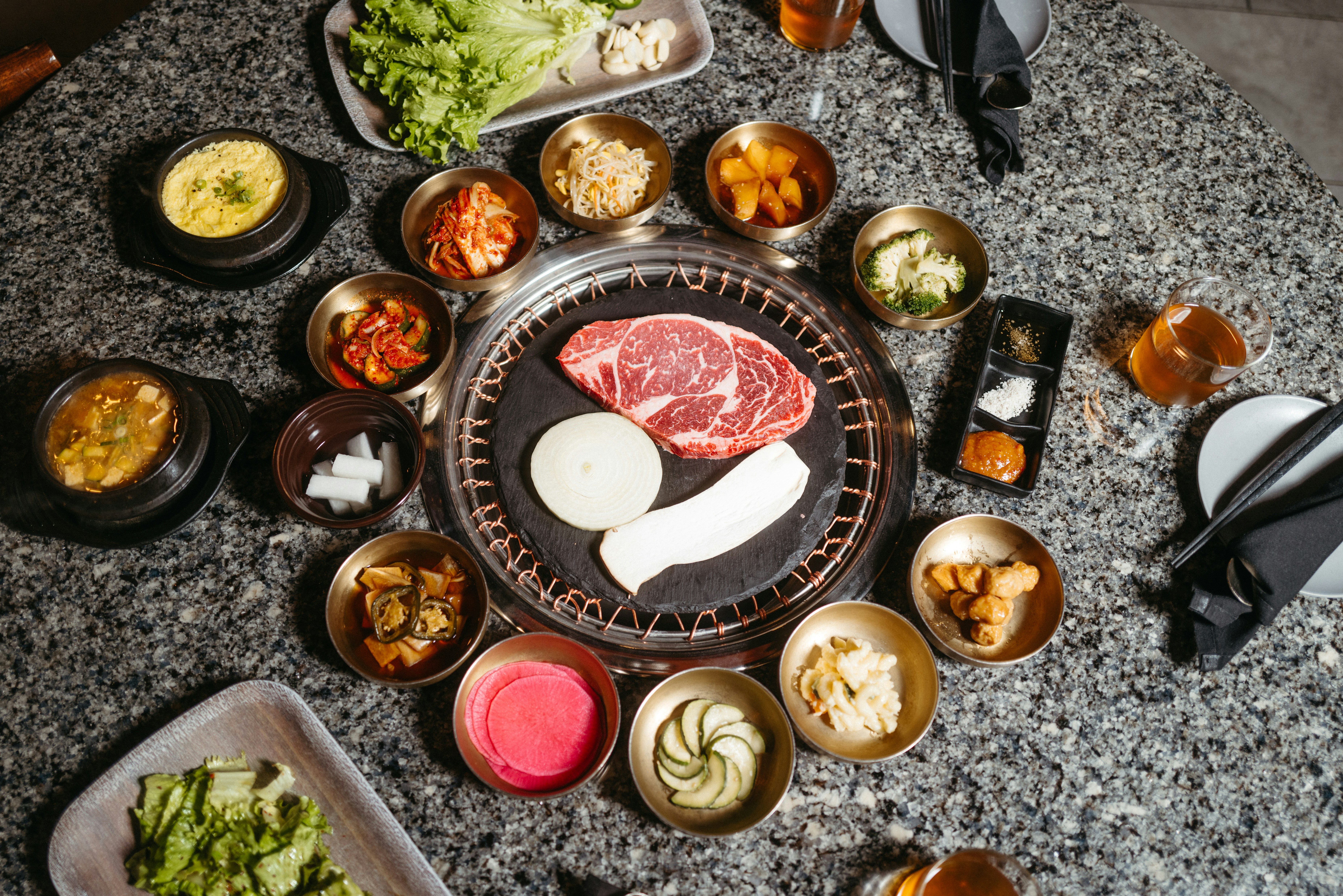 a table topped with lots of different types of food, An enticing display of Korean BBQ ingredients, from thinly sliced beef to crisp lettuce wraps, inviting you to create your perfect bite.