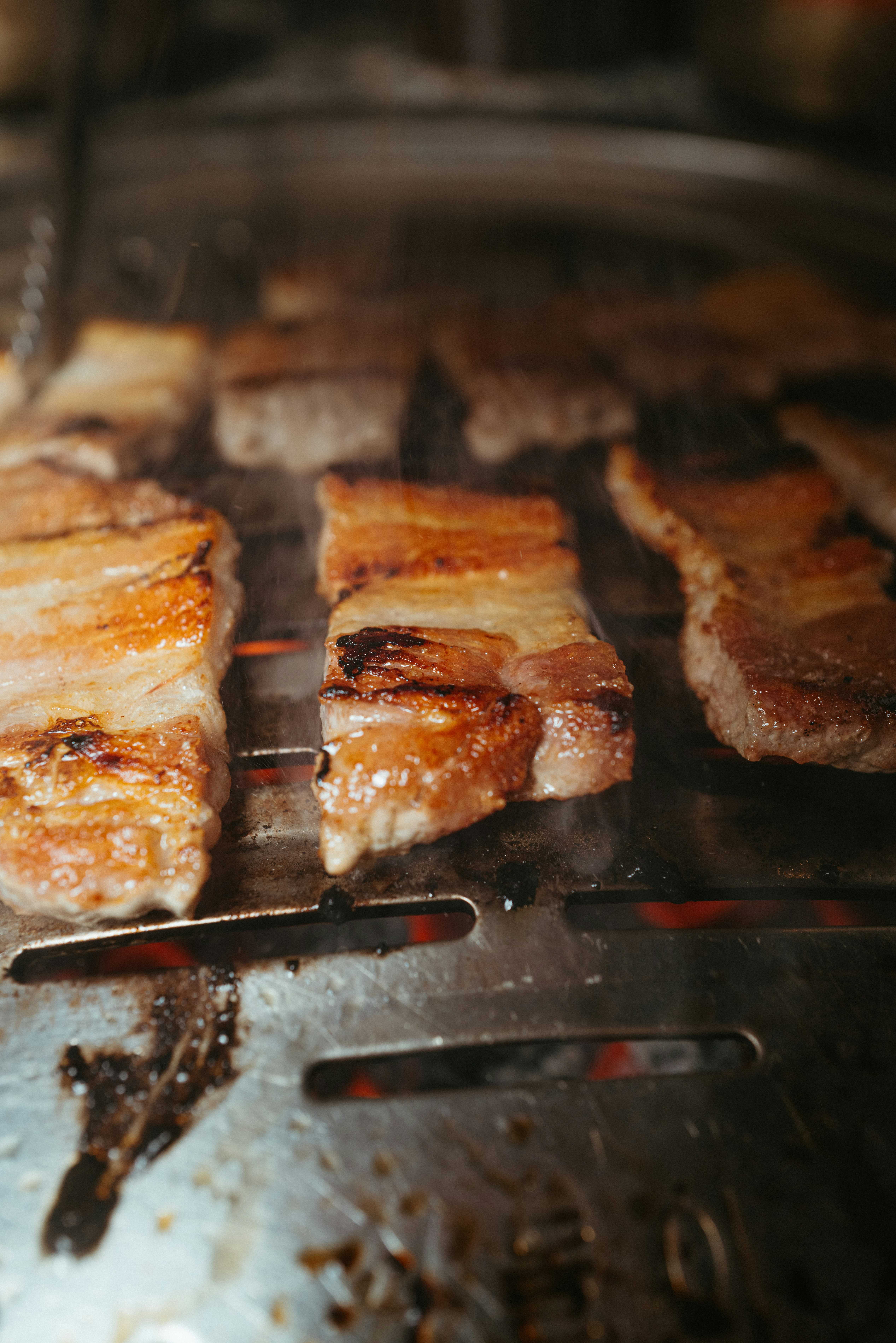 Grilled pork belly slices on a grill grate with char marks, smoky atmosphere, warm orange firelight