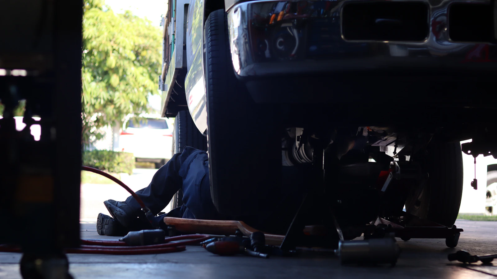 Technician lying on a creeper beneath a raised pickup truck, working on the underside in a repair bay.