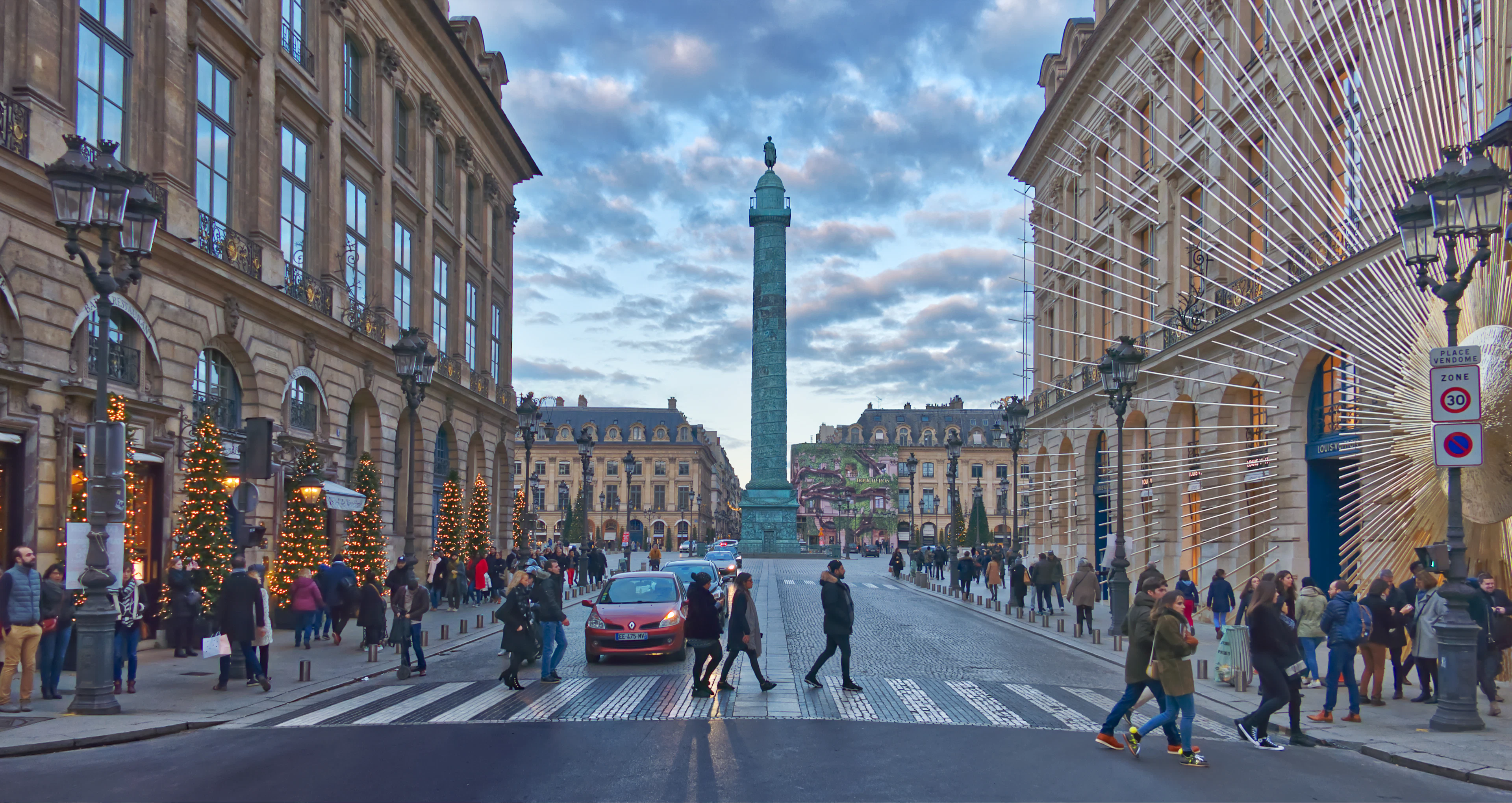Pedestrians cross Place Vendôme at dusk, Christmas lights strung along the shopfronts.