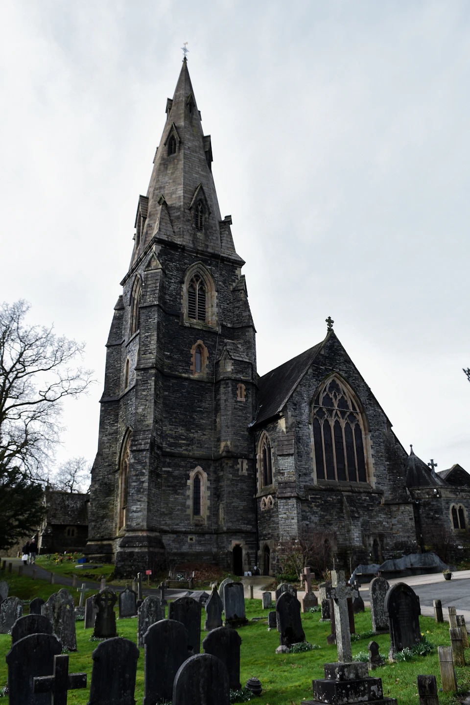 The image depicts a large, Gothic-style church with a tall spire and multiple levels, standing majestically against an overcast sky. The church is surrounded by several gravestones, indicating its historical significance. In front of the church, there are numerous black headstones arranged in rows, each one bearing the names of those who passed away.

The perspective of the image is from a low angle, looking up at the church and the gravestones, which enhances the imposing presence of the structure. The overcast sky casts a soft light on the scene, highlighting the intricate details of the church's architecture and the solemnity of its surroundings.