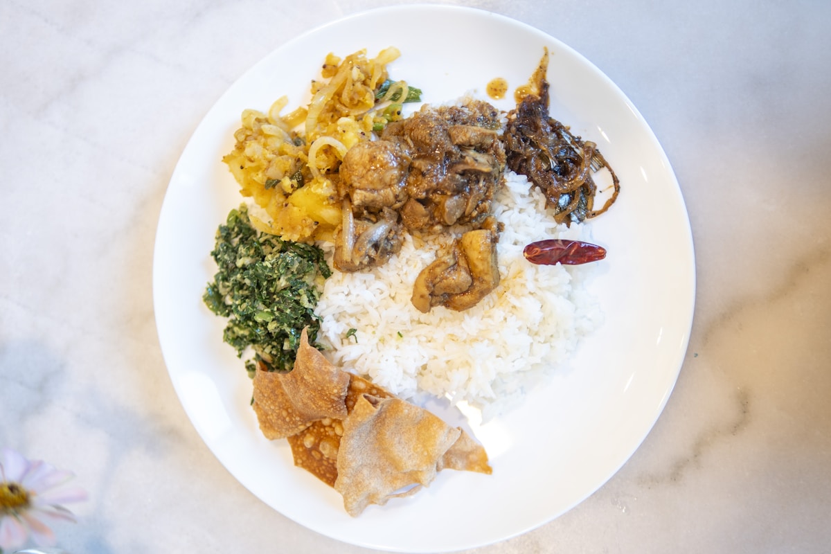 A traditional Sri Lankan rice and curry plate with multiple small dishes arranged around white rice - dhal, green leaves, potato deviled, and papadam