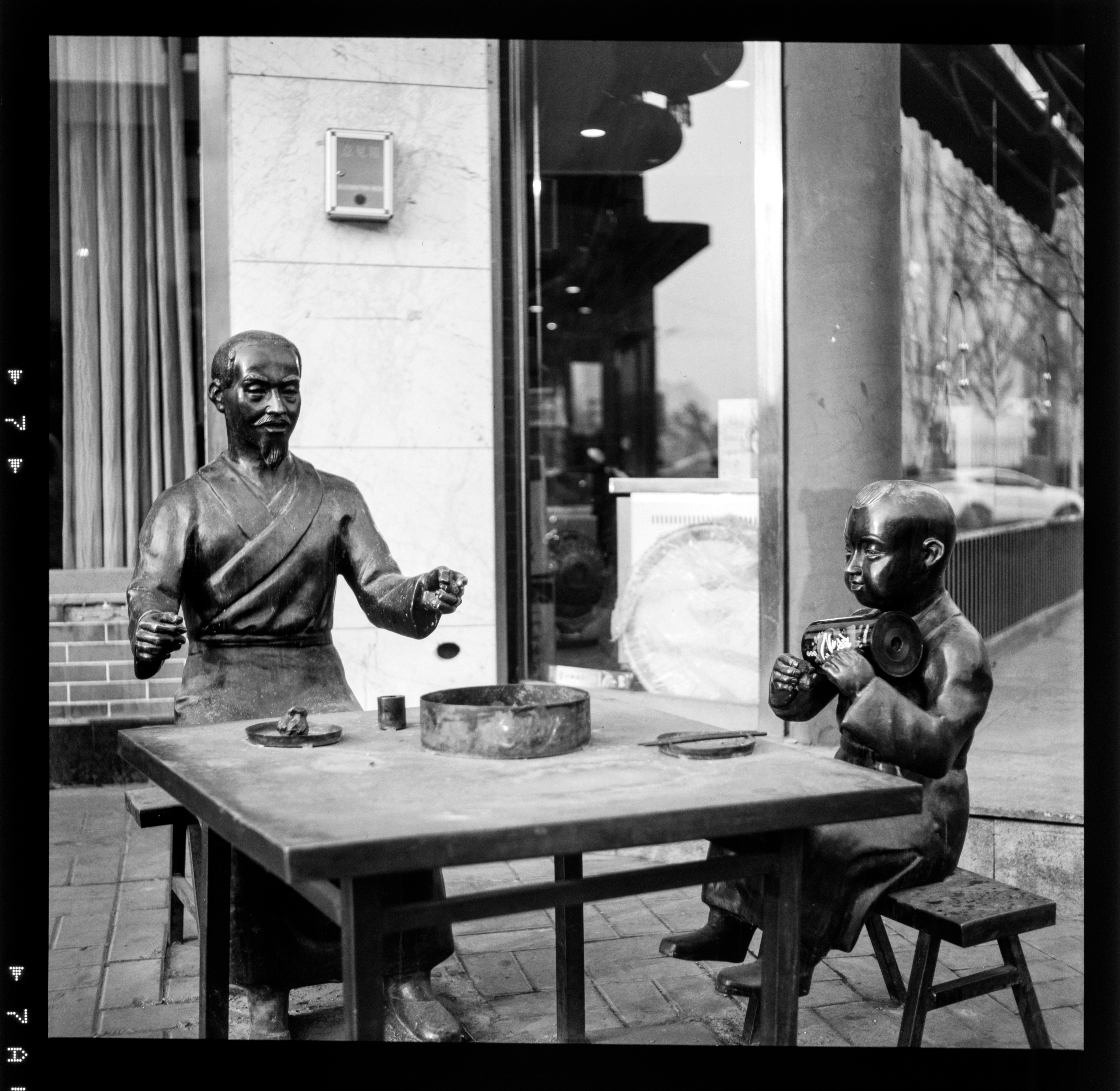 Two life-sized bronze figures sit at a rough wooden table in an urban storefront setting, one gesturing while the other studies a small object; the scene feels like a street-side diorama or sculpture display.