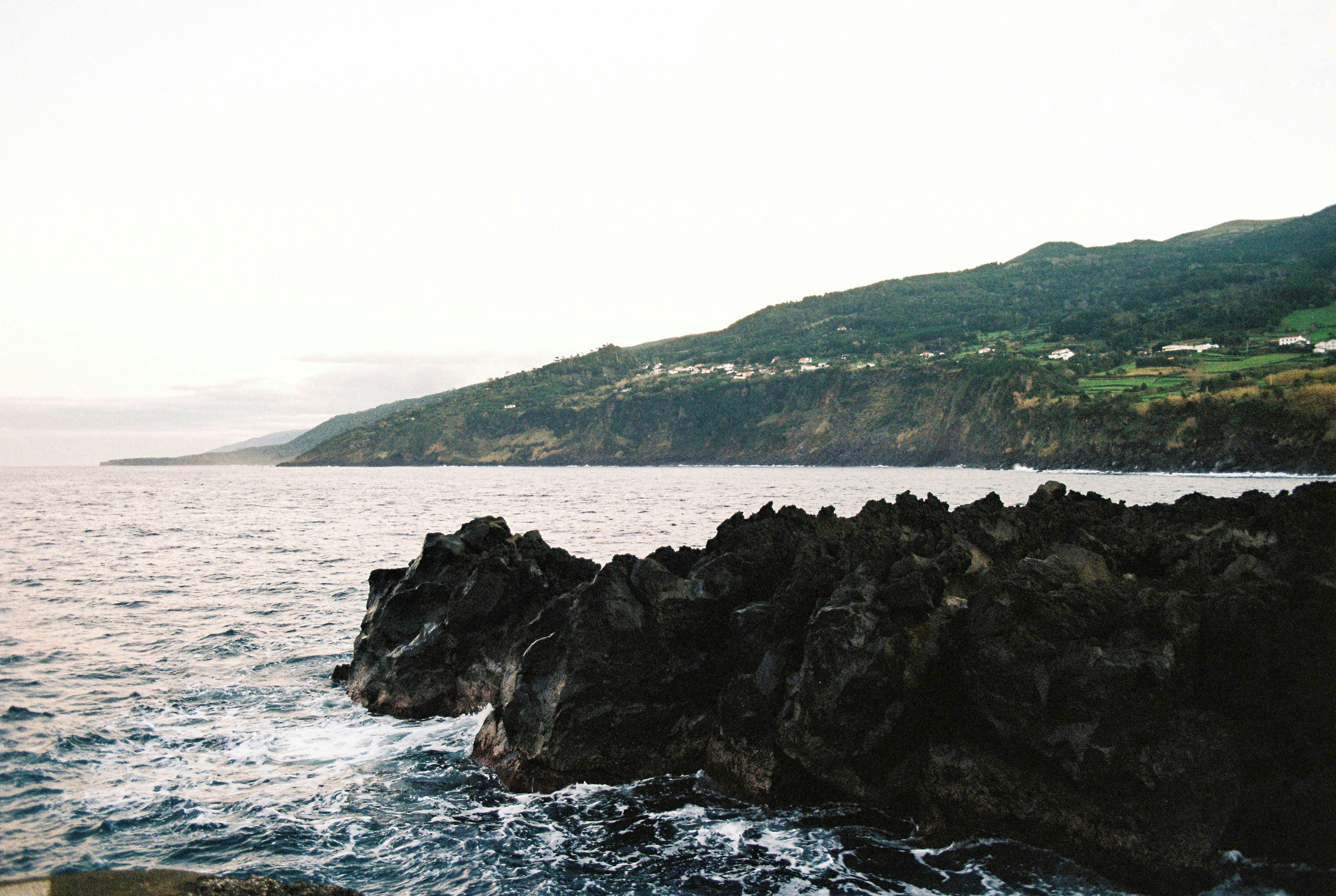 a view of a body of water with mountains in the background, The sea crashing against the rocks on Pico Island, Azores on film.
