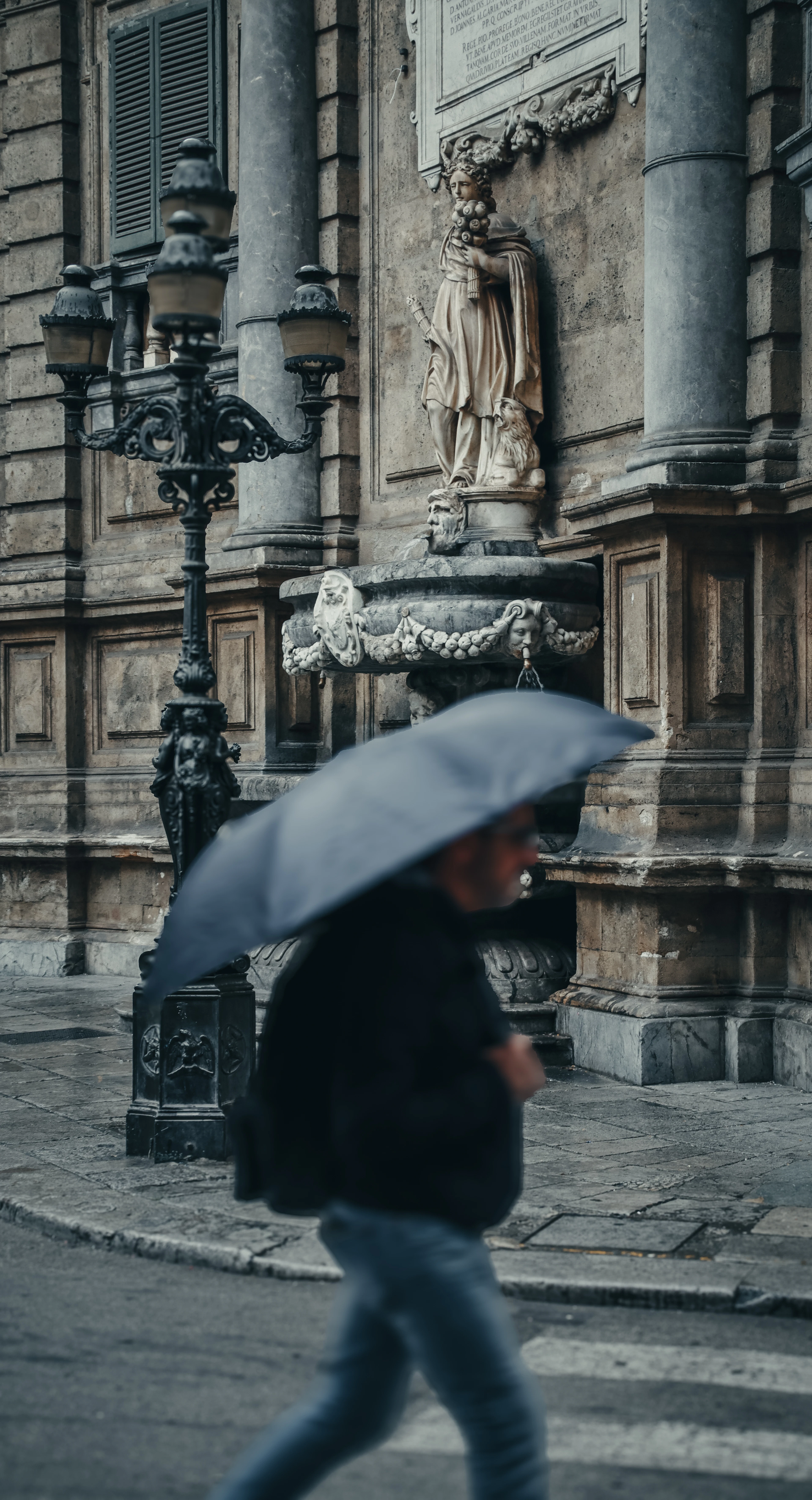 A pedestrian with an umbrella passes a baroque statue niche and wrought-iron lamppost on a Palermo street corner.