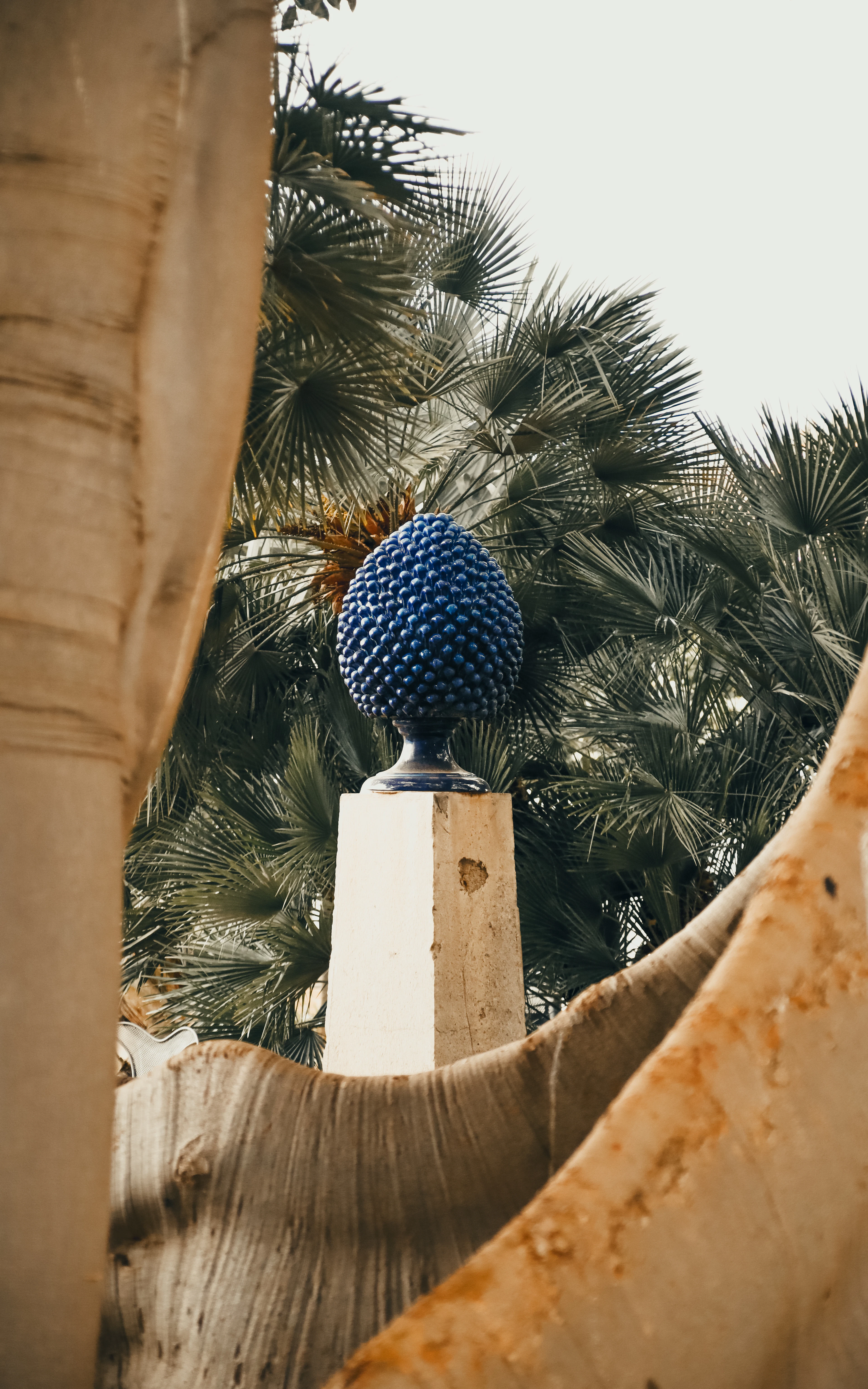 A blue ceramic pigna (pinecone) finial on a stone pillar, framed by palm fronds in a Palermo garden.