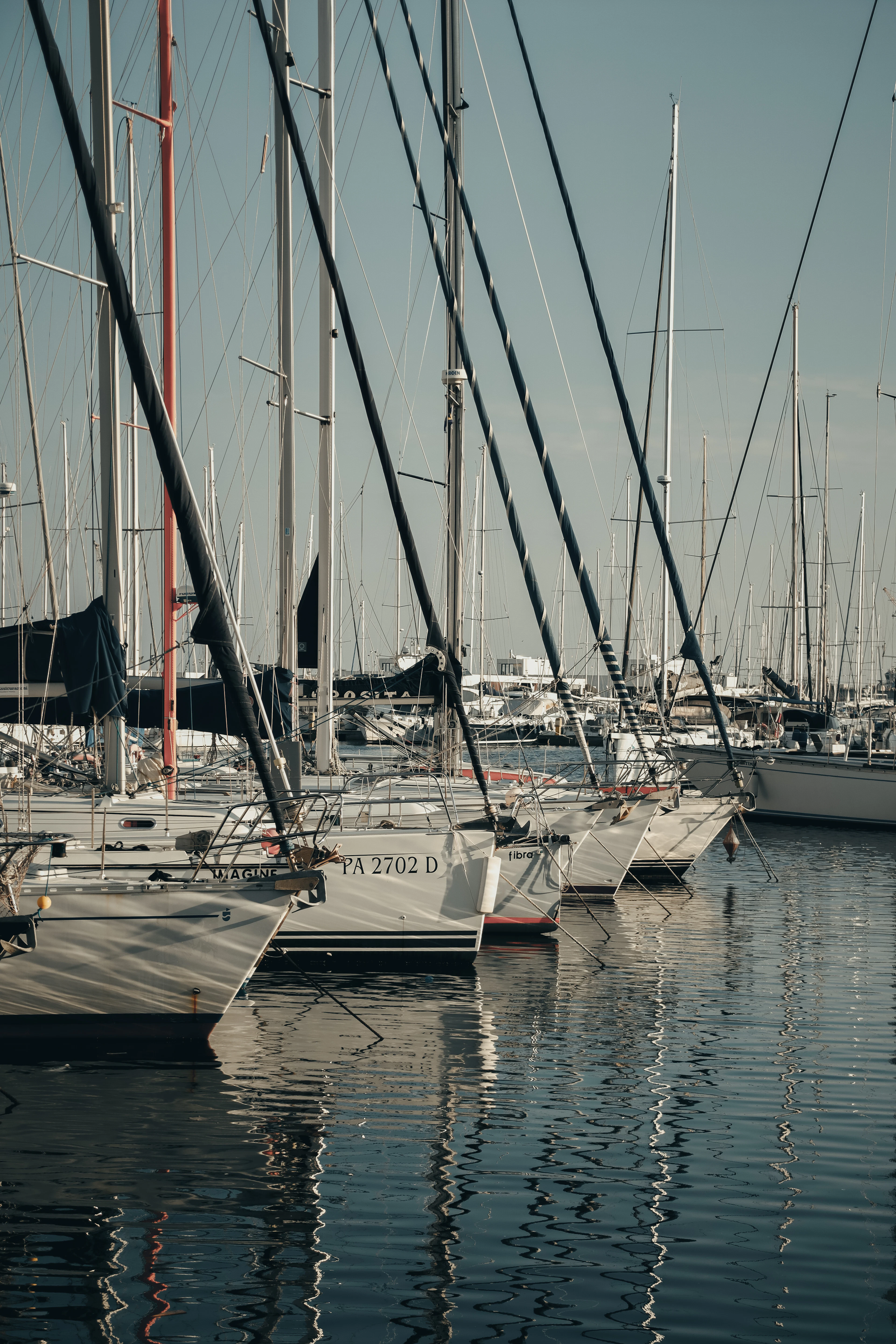 Sailboats moored in a Palermo marina, their masts and rigging reflected in calm water.