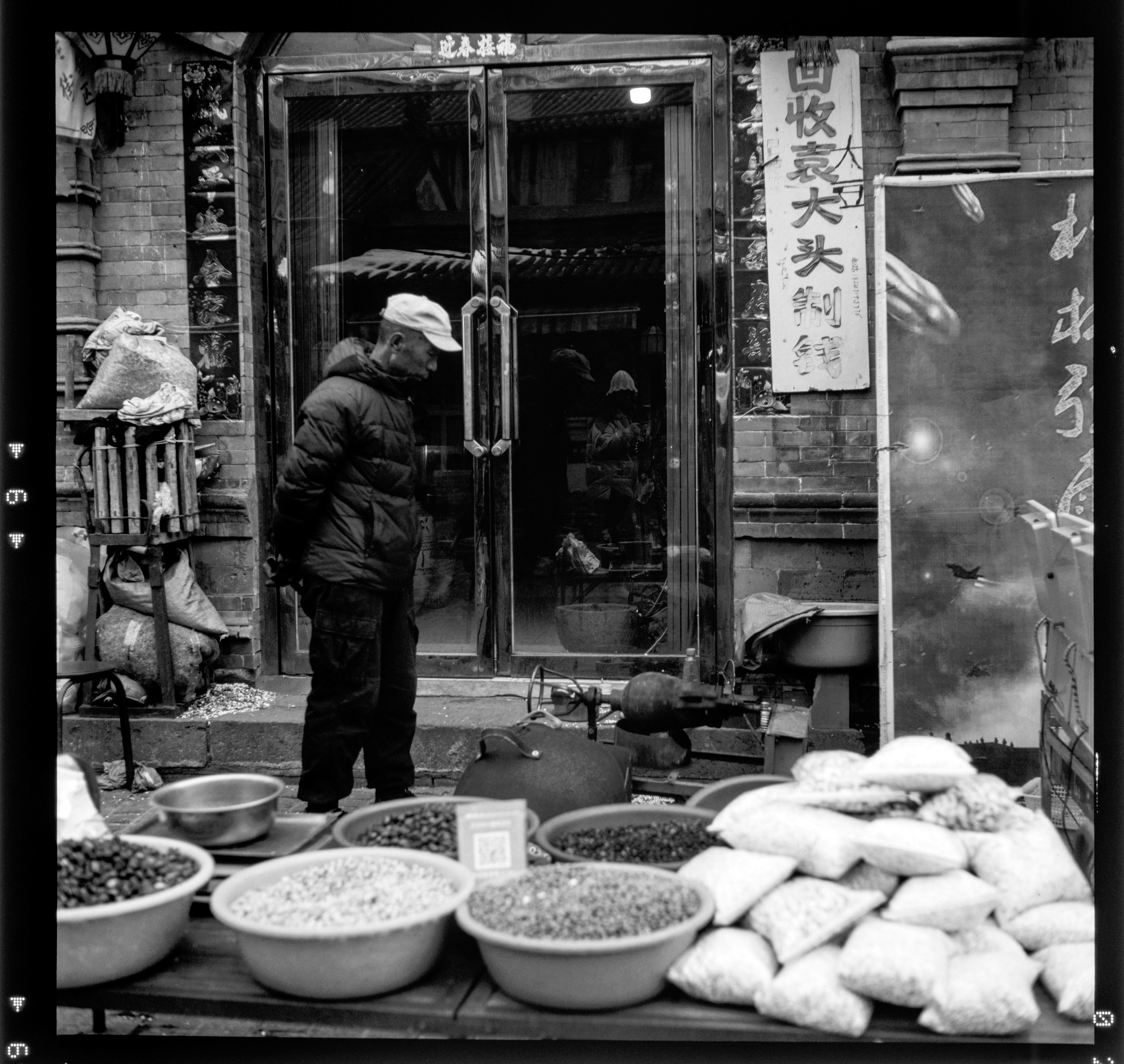 Black-and-white street-market scene with a man in a puffer jacket near a shop doorway, foreground bowls of grains and stacked sacks.