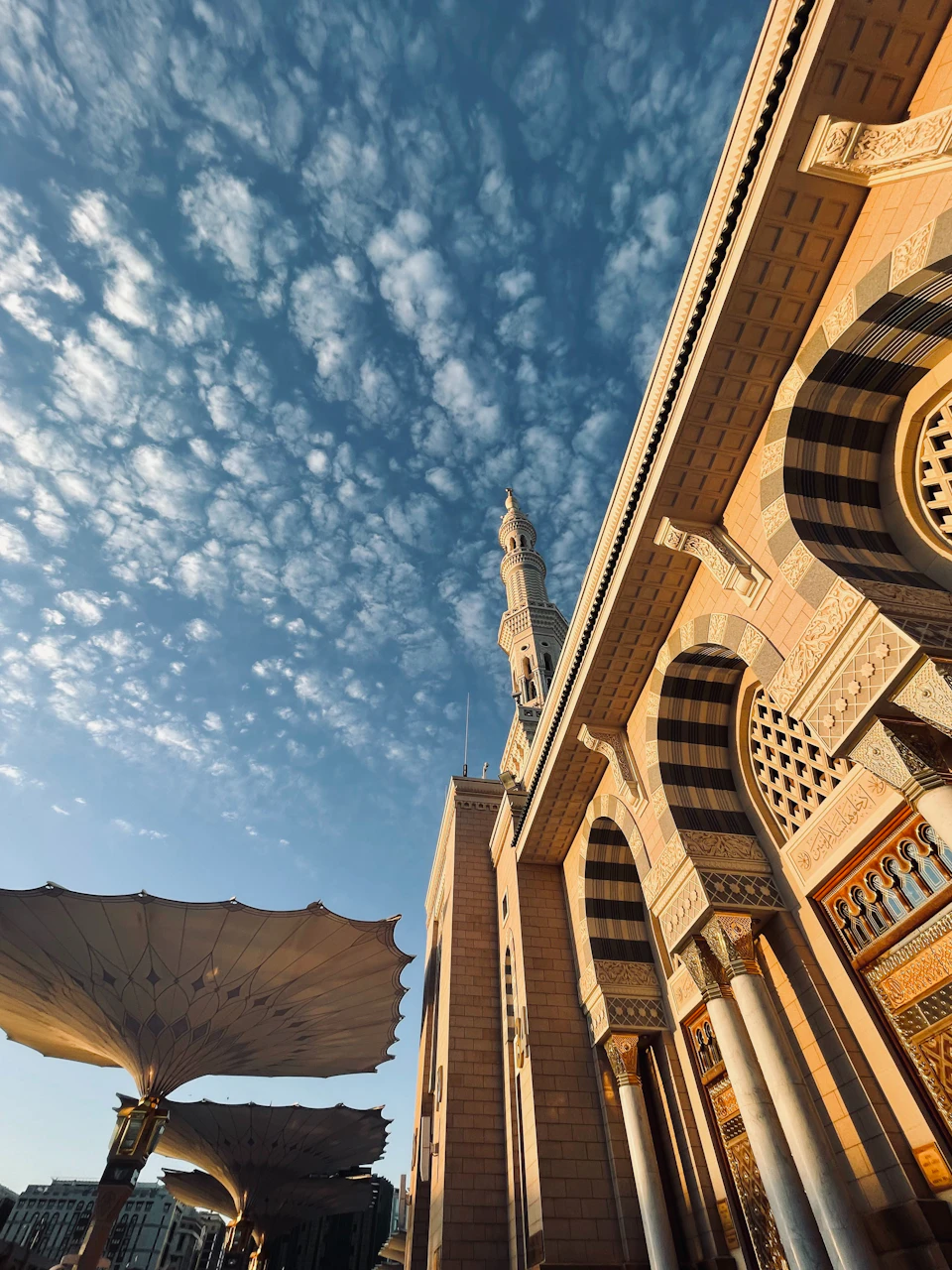 The image depicts a large, ornate building with intricate architectural details and a prominent minaret reaching towards the sky. The building is bathed in warm sunlight, highlighting its grandeur against the backdrop of a clear blue sky dotted with fluffy white clouds. In front of the building, there are two large umbrellas providing shade to the area around them.