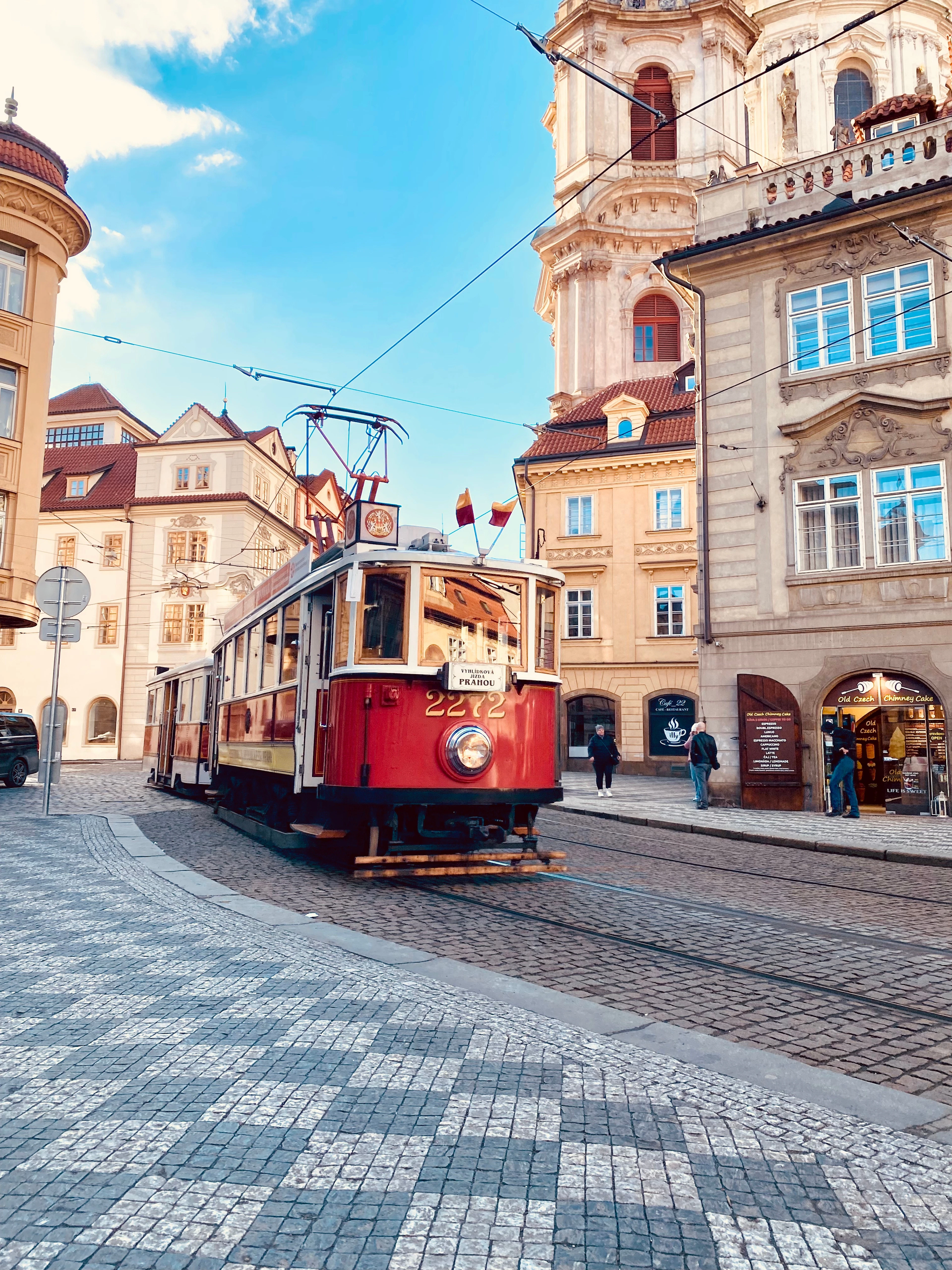 A historic red tram turning across cobblestones in Prague's Mala Strana, baroque facades and a bell tower behind