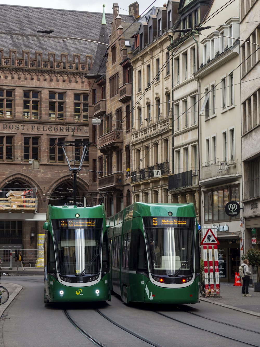 a couple of green trains traveling down a street next to tall buildings