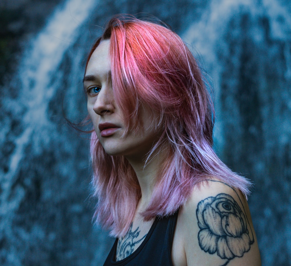 a woman with pink hair and piercings standing in front of a waterfall