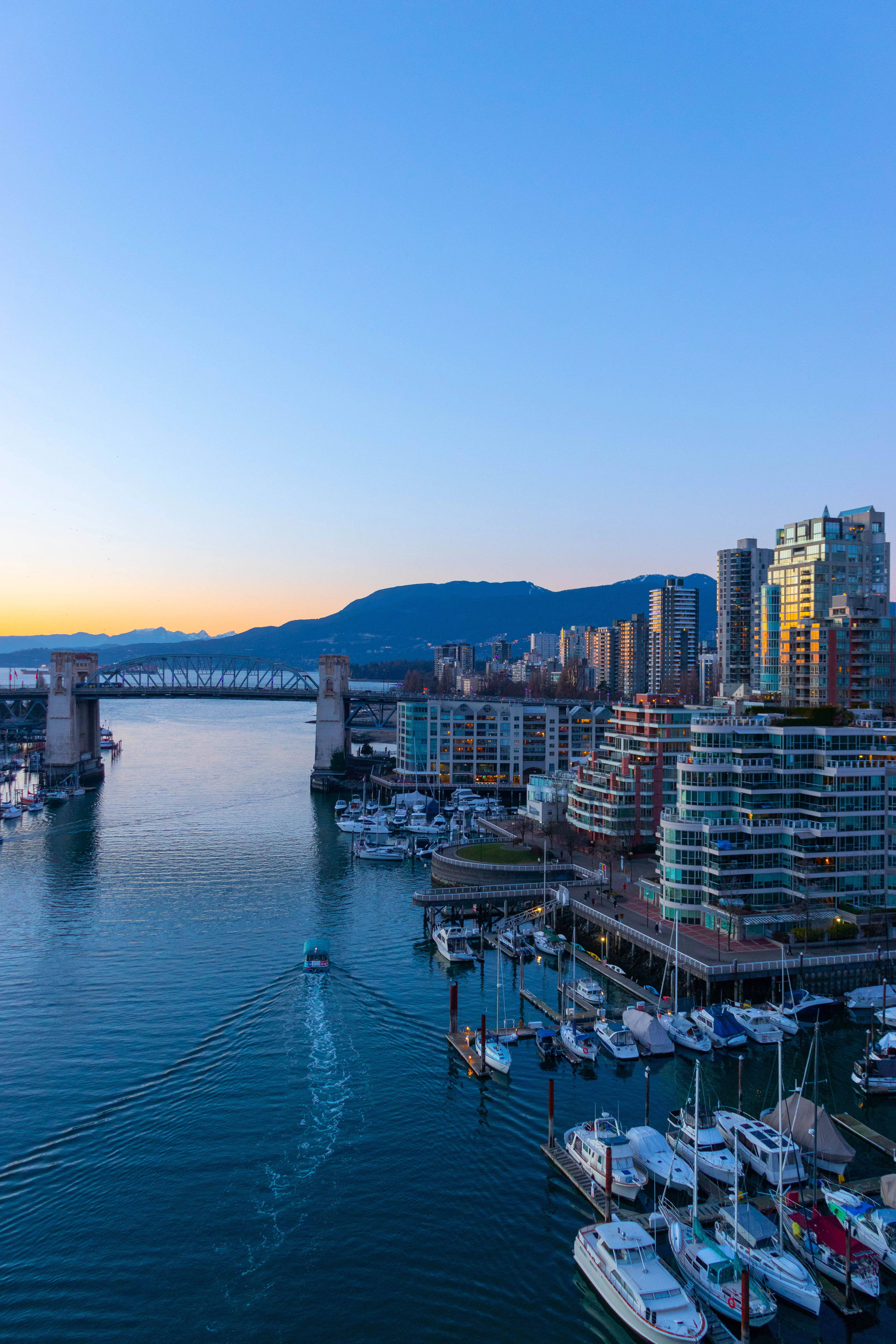 Sailboats and yachts moored in False Creek marina at dusk, glowing high-rises and an old railway bridge behind.