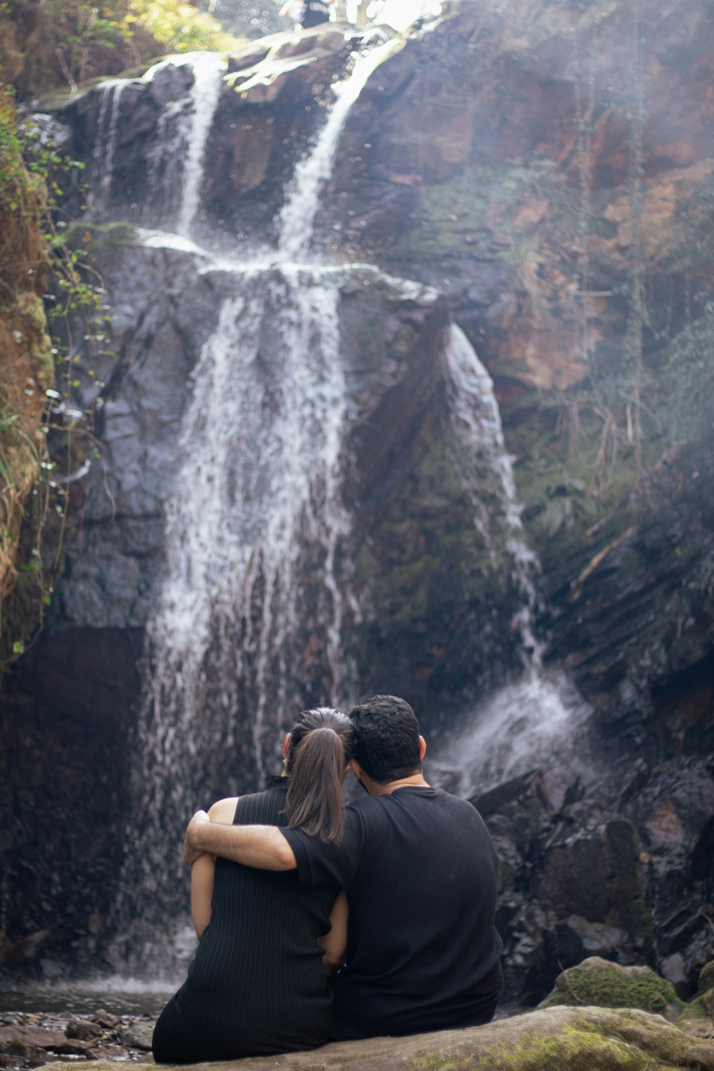 a man and woman sitting on a rock in front of a waterfall