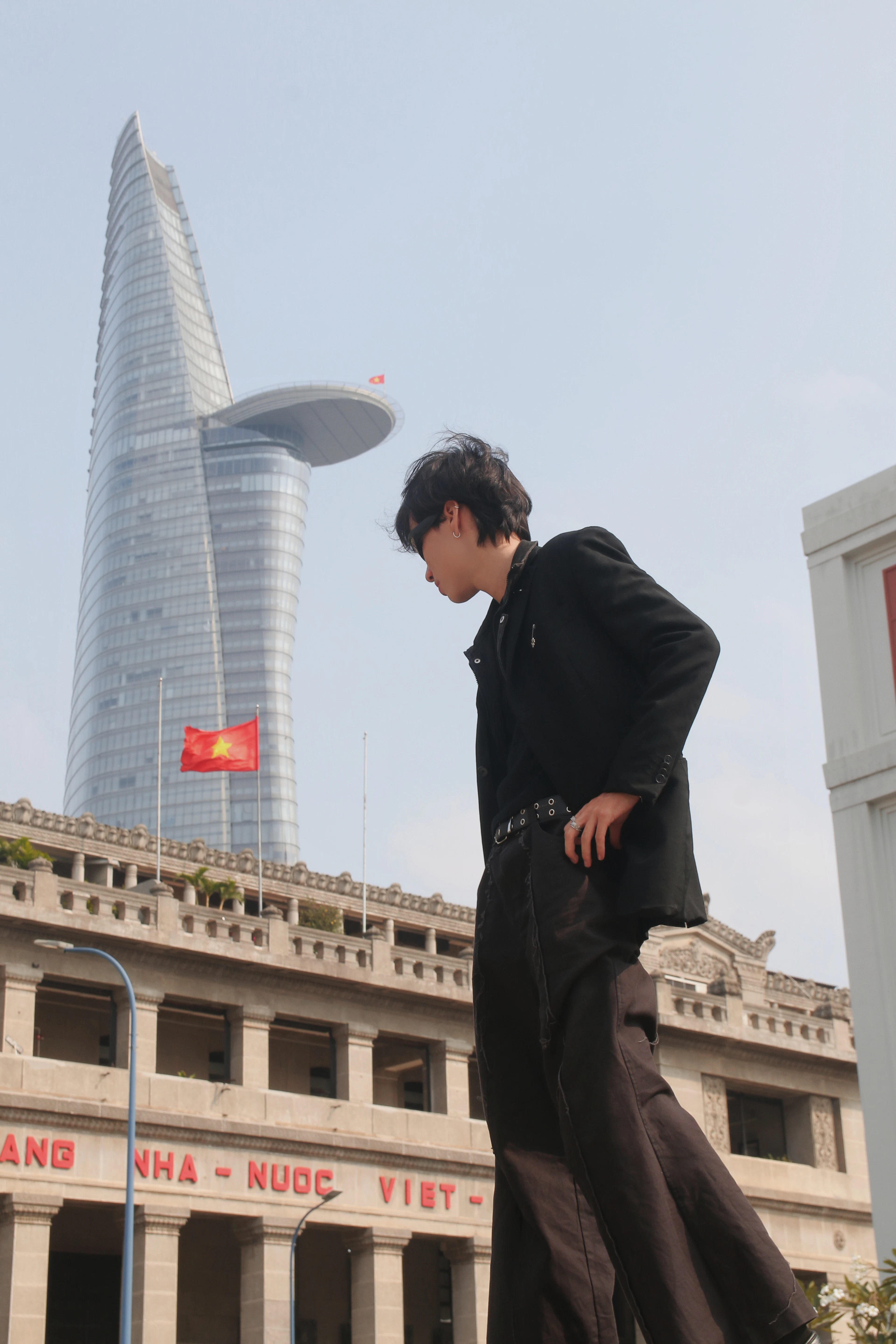 A person in black poses on a street in Ho Chi Minh City with the spiral-topped Bitexco Financial Tower and a Vietnamese flag behind them