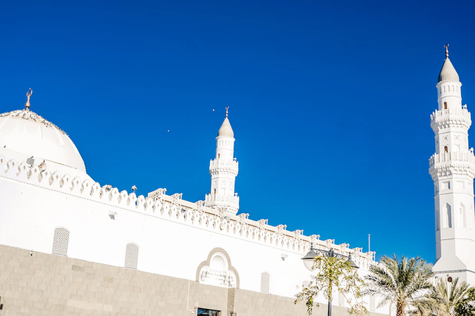 The image depicts a serene scene at a mosque, with two prominent white minarets reaching towards the clear blue sky. The minarets are adorned with small spires and domes, adding to their majestic appearance. In front of these structures is a large white wall, which features arched windows that allow natural light to filter into the space.

In addition to the mosque's architectural elements, there are several palm trees scattered throughout the scene, providing a touch of nature amidst the man-made structures. The image captures the beauty and tranquility of this religious site against the backdrop of a bright blue sky.