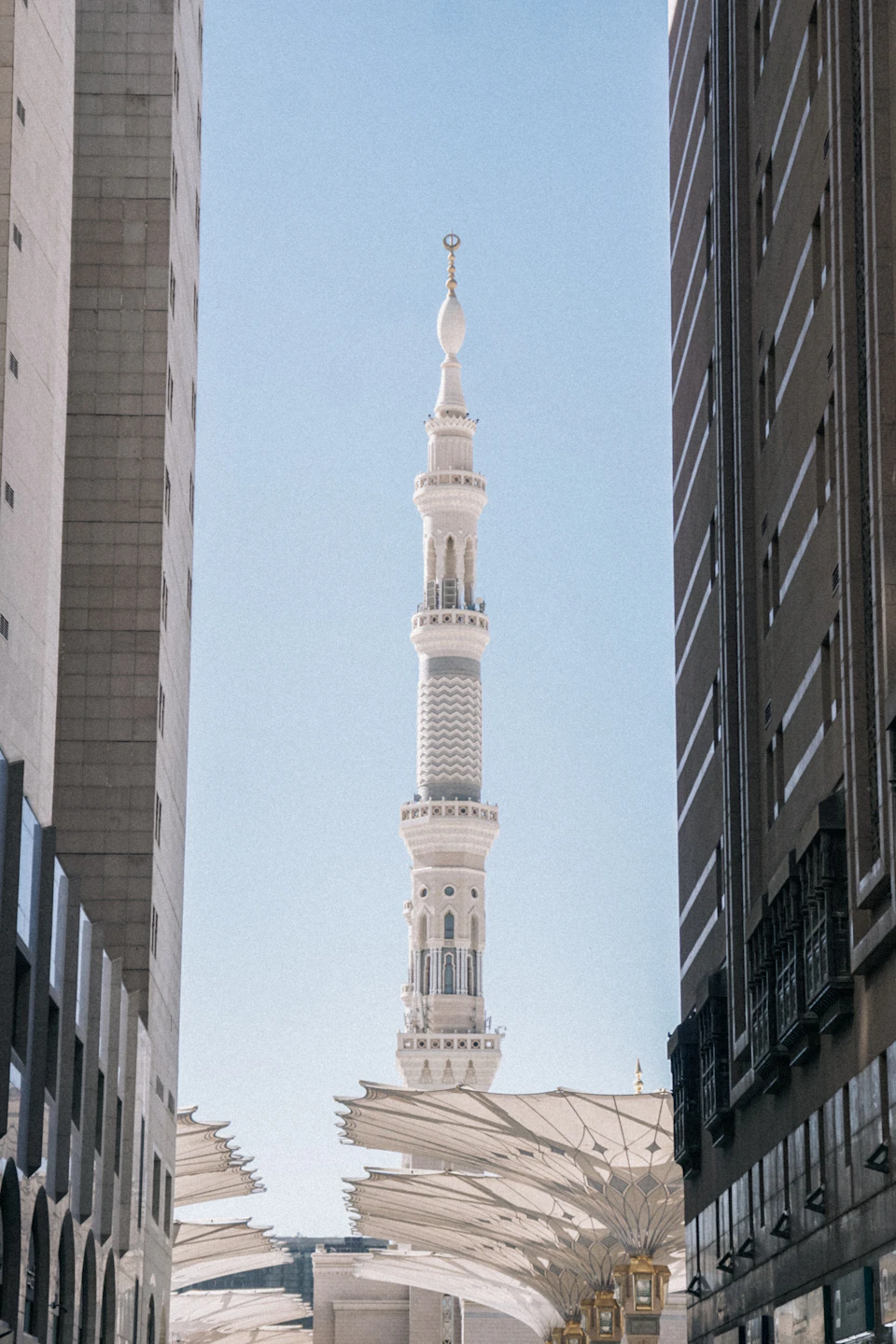 The image shows a tall, white mosque with multiple levels and a pointed spire at its peak. The mosque is surrounded by modern buildings on either side, creating an interesting contrast between traditional architecture and contemporary structures. The sky above is clear blue, providing a serene backdrop to the scene.
