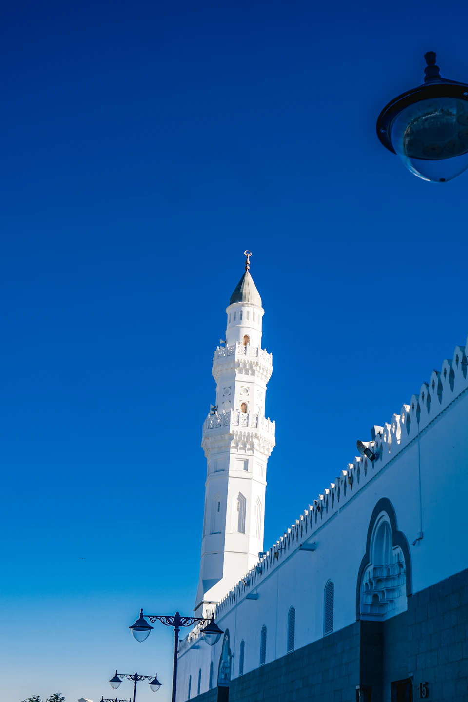 The image depicts a majestic white mosque with a tall minaret and a dome, standing against a clear blue sky. The mosque is situated on a street corner, surrounded by other buildings that create an urban backdrop to its architectural grandeur. A lamppost can be seen in the foreground of the photo, adding depth to the scene.