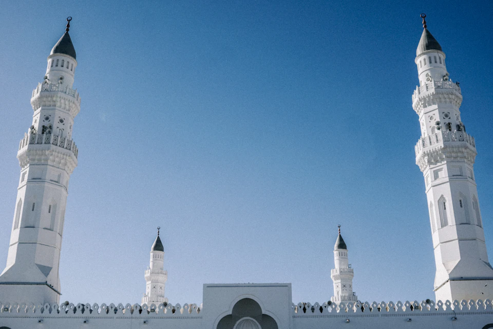 The image depicts a majestic white mosque with four towering minarets, each adorned with black spires that reach towards the clear blue sky. The mosque is situated on a hillside, providing an elevated perspective for its viewers. The photograph captures the intricate details of the architectural design and the grandeur of the structure.