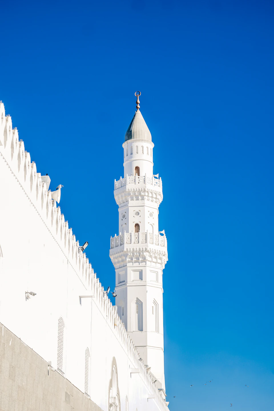 The image depicts a majestic white mosque with multiple levels, including a tall minaret and a smaller tower on each level. The mosque is situated against a clear blue sky, creating a striking contrast between the white structure and the vibrant backdrop. A few birds can be seen flying in the background, adding to the serene atmosphere of the scene.

The image does not contain any discernible text or human figures, making it an ideal representation of a mosque's architectural design. The focus is solely on the mosque itself, highlighting its grandeur and beauty against the backdrop of nature.