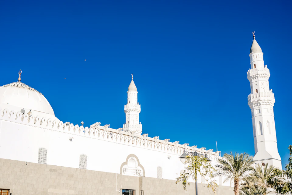 The image depicts a serene scene at a mosque, with two prominent white minarets reaching towards the clear blue sky. The minarets are adorned with small domes and topped with spires that add to their majestic appearance. In front of these minarets is a large white wall, which features an intricate design on its surface.

In addition to the minarets, there are two palm trees visible in the foreground, adding a touch of nature to the scene. The image does not contain any discernible text or countable objects, and the relative positions of the elements suggest that they are stationary within their respective environments.