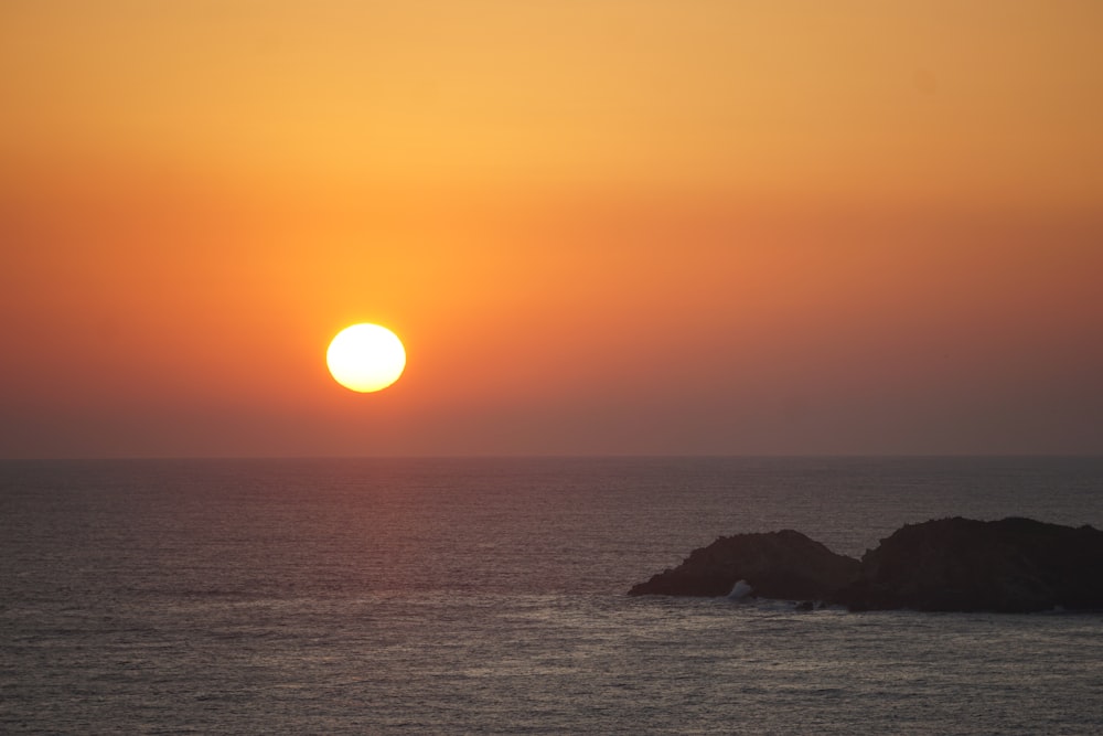 the sun is setting over the ocean with rocks in the foreground