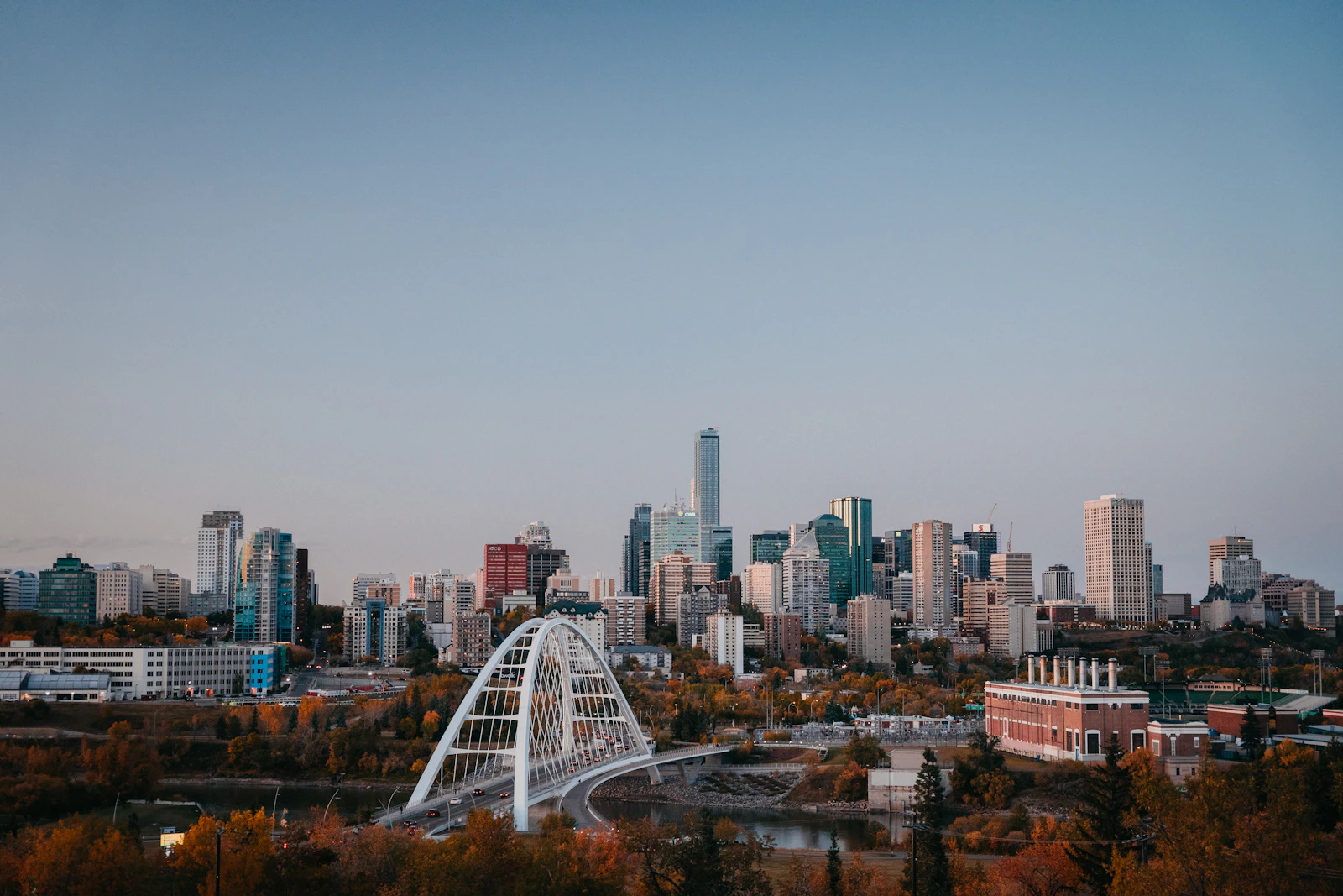 The Edmonton skyline at sunset across the river valley with the white arch of the Walterdale Bridge and autumn trees in the foreground