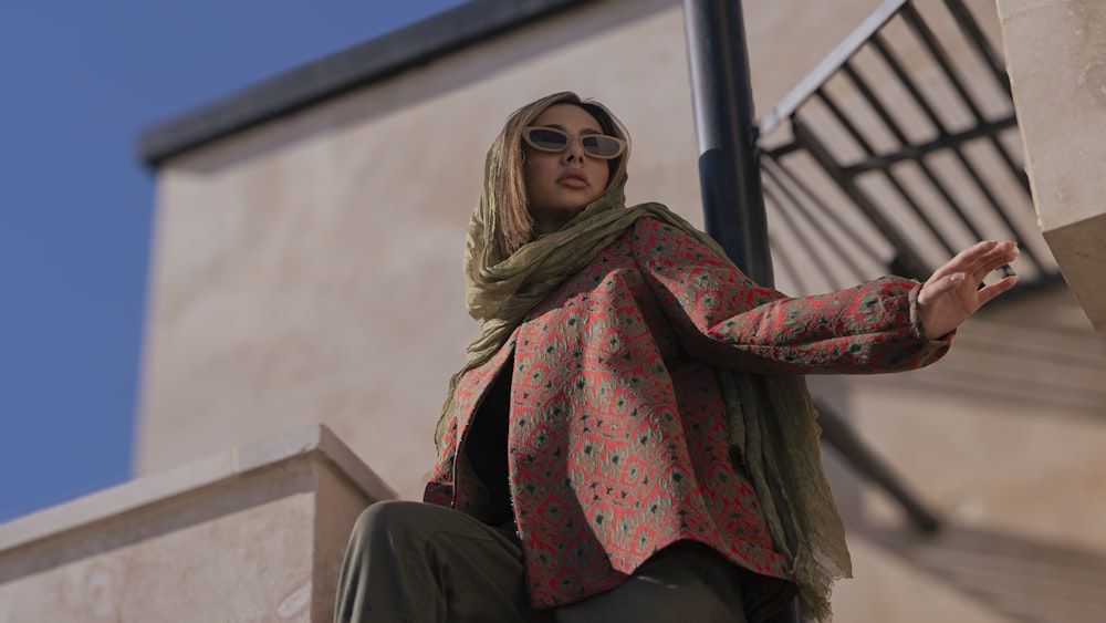 a woman wearing a scarf and sunglasses sitting on a ledge