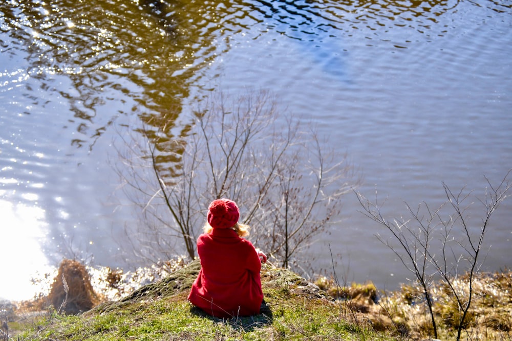 a little girl in a red jacket sitting on a hill looking at a pond