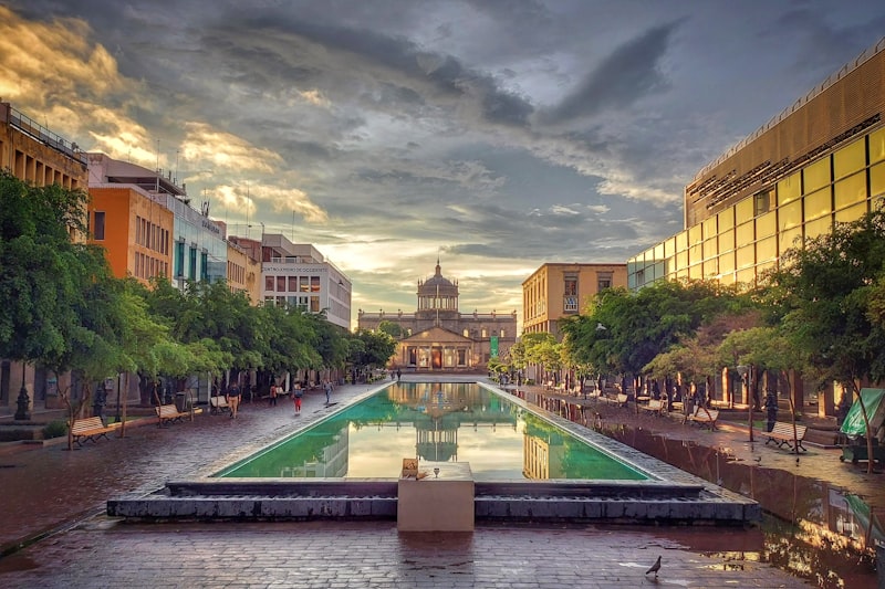 A long reflecting pool on Plaza Tapatía at sunrise, leading toward the domed Hospicio Cabañas between rows of trees