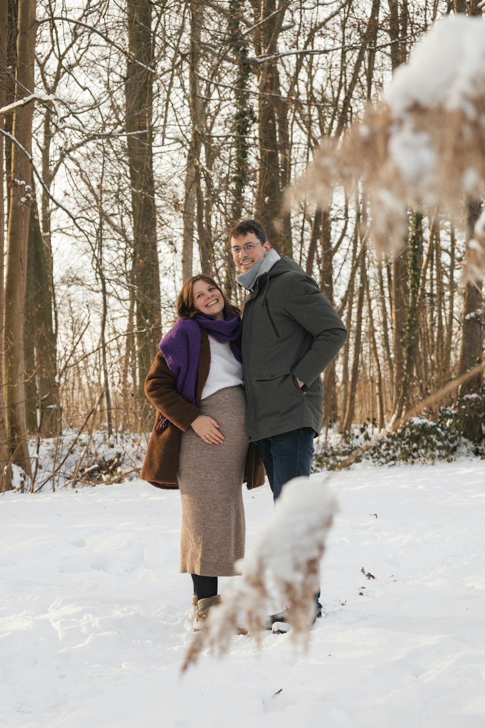 a pregnant couple standing in the snow in the woods