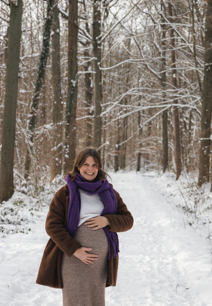 A pregnant woman in a brown coat and purple scarf walking on a snowy forest path