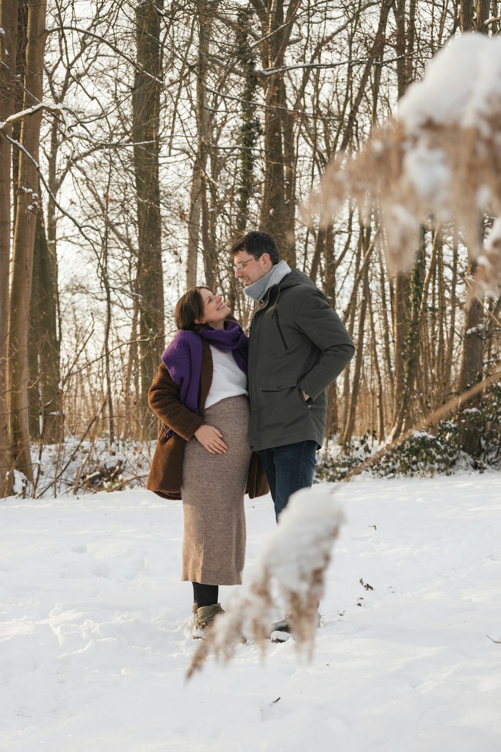 a pregnant couple standing in the snow in a wooded area