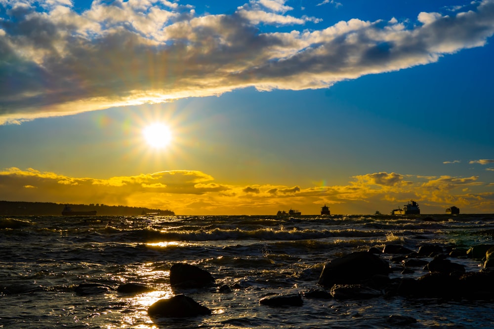 the sun is setting over the ocean with rocks in the foreground