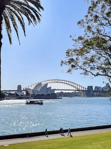 people walking on a path near the water with a bridge in the background