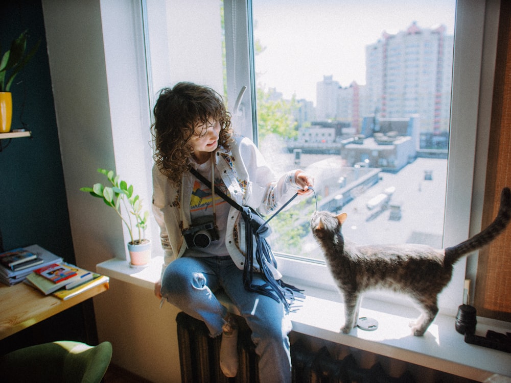 a woman sitting on a window sill petting a cat