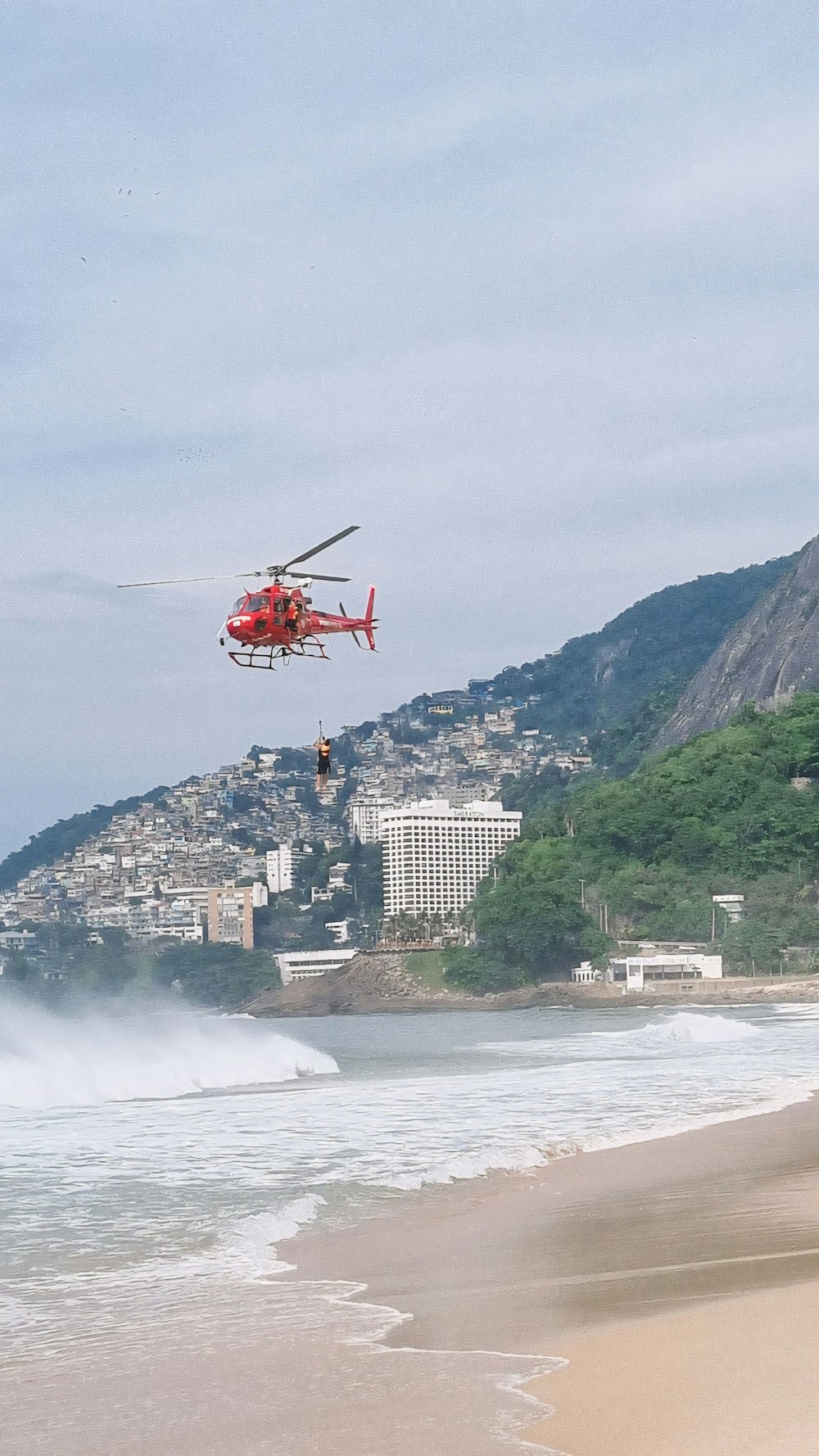 a red helicopter flying over a beach next to the ocean